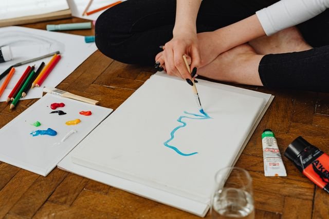 Close-up of a person painting on paper, surrounded by colorful art supplies on a wooden floor.